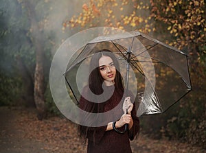 Young pensive woman under a transparent umbrella on a background of autumn and smoke.