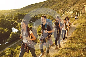Young people with backpacks walking along narrow path in mountains. Group of cheerful tourists hiking outside in summer