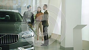 Young people buying car in motor showroom talking to salesperson looking at automobile