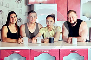 Young people drink hot drinks while sitting at kitchen island, two couples in their 20s look at camera in country house.