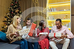 Young people friends give each other and receive New Year present gift box. Christmas eve sitting at home on the couch