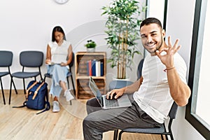 Young people sitting at waiting room working with laptop doing ok sign with fingers, smiling friendly gesturing excellent symbol