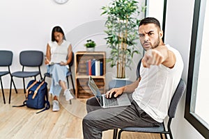 Young people sitting at waiting room working with laptop pointing with finger to the camera and to you, confident gesture looking