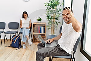 Young people sitting at waiting room working with laptop smiling happy doing ok sign with hand on eye looking through fingers