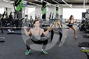 Young people working out with barbells at gym. Attractive women and handsome muscular man trainer are training in light