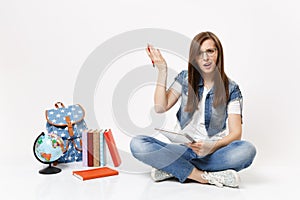 Young perplexed woman student in glasses spreading hands holding pencil and notebook sitting near globe backpack books