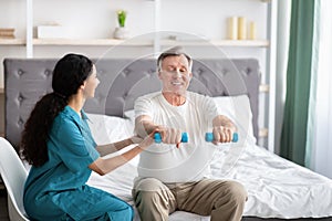 Young physiotherapist helping homebound elderly man to do physical exercises with dumbbells on bed at home photo