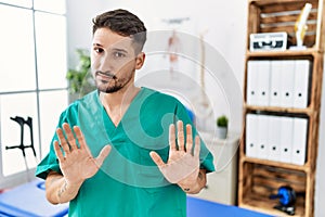 Young physiotherapist man working at pain recovery clinic moving away hands palms showing refusal and denial with afraid and