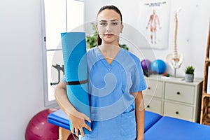 Young physiotherapist woman holding yoga mat thinking attitude and sober expression looking self confident