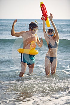 Young playful couple on the beach in water playing with water gun. Woman feeling victorius, man feeling defeated. Holiday,