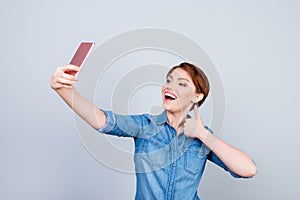 Young playful girl is making selfie on the camera of her telephone and showing like sign