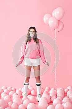 Young playful woman blowing bubble with chewing gum staying in pink air balloons background