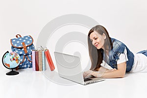 Young pleasant woman student in denim clothes working on laptop pc computer lying near globe, backpack, school books