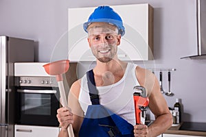 Young Plumber Holding Wrench And Plunger In Kitchen