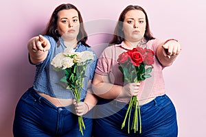 Young plus size twins holding flowers pointing with finger to the camera and to you, confident gesture looking serious