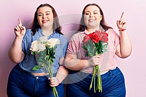 Young plus size twins holding flowers surprised with an idea or question pointing finger with happy face, number one