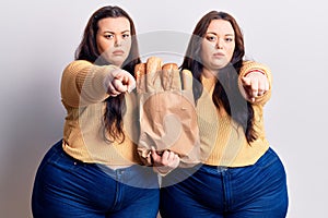 Young plus size twins holding paper bag with bread pointing with finger to the camera and to you, confident gesture looking