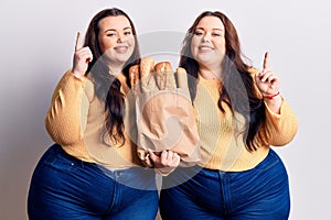 Young plus size twins holding paper bag with bread surprised with an idea or question pointing finger with happy face, number one