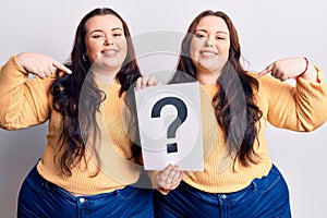 Young plus size twins holding question mark pointing finger to one self smiling happy and proud