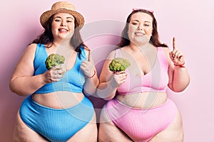 Young plus size twins wearing bikini holding broccoli smiling with an idea or question pointing finger with happy face, number one