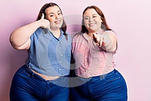 Young plus size twins wearing casual clothes smiling doing talking on the telephone gesture and pointing to you