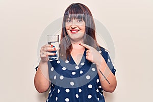 Young plus size woman drinking glass of water smiling happy pointing with hand and finger