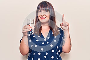 Young plus size woman drinking glass of water smiling with an idea or question pointing finger with happy face, number one
