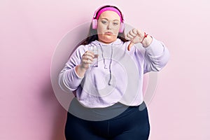 Young plus size woman drinking glass of water using headphones with angry face, negative sign showing dislike with thumbs down,