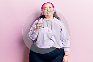 Young plus size woman drinking glass of water using headphones looking positive and happy standing and smiling with a confident