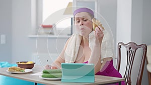 Young plus-size woman eating sandwich writing calories looking at tablet screen. Portrait of serious Caucasian obese