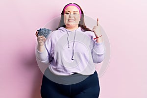 Young plus size woman holding blueberries smiling with an idea or question pointing finger with happy face, number one