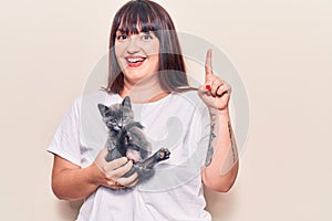 Young plus size woman holding cat smiling with an idea or question pointing finger with happy face, number one