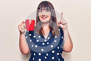 Young plus size woman holding coffee smiling with an idea or question pointing finger with happy face, number one