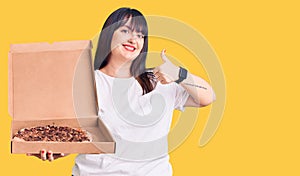Young plus size woman holding delivery pizza box smiling happy and positive, thumb up doing excellent and approval sign