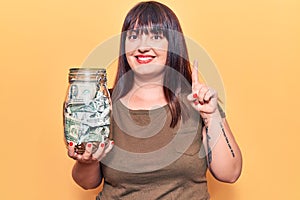 Young plus size woman holding jar with savings smiling with an idea or question pointing finger with happy face, number one