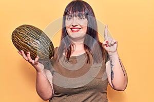 Young plus size woman holding melon smiling with an idea or question pointing finger with happy face, number one