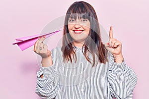 Young plus size woman holding paper airplane smiling with an idea or question pointing finger with happy face, number one