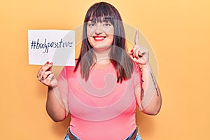 Young plus size woman holding paper with hashtag body positive smiling with an idea or question pointing finger with happy face,