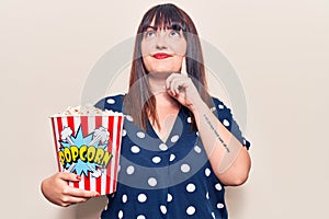 Young plus size woman holding popcorn serious face thinking about question with hand on chin, thoughtful about confusing idea