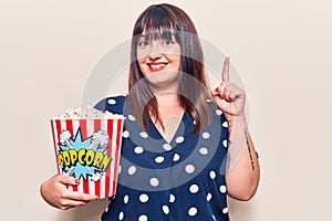 Young plus size woman holding popcorn surprised with an idea or question pointing finger with happy face, number one