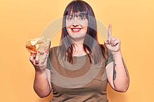 Young plus size woman holding potato chip surprised with an idea or question pointing finger with happy face, number one