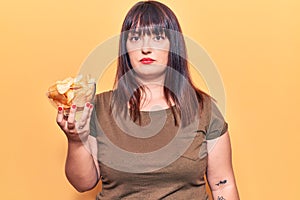 Young plus size woman holding potato chip thinking attitude and sober expression looking self confident