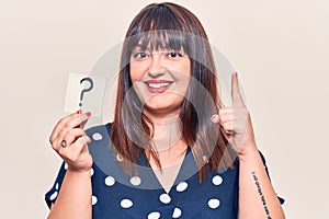 Young plus size woman holding question mark smiling with an idea or question pointing finger with happy face, number one