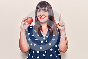 Young plus size woman holding red apple smiling with an idea or question pointing finger with happy face, number one