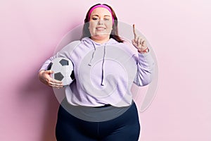 Young plus size woman holding soccer ball smiling with an idea or question pointing finger with happy face, number one