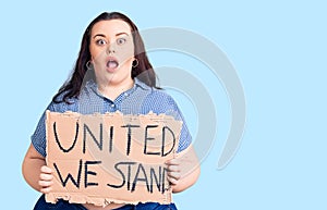 Young plus size woman holding united we stand banner scared and amazed with open mouth for surprise, disbelief face