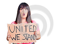 Young plus size woman holding united we stand banner scared and amazed with open mouth for surprise, disbelief face