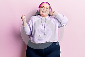 Young plus size woman listening to music using headphones screaming proud, celebrating victory and success very excited with