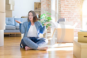 Young plus size woman sitting on the floor around cardboard boxes moving to a new home clueless and confused expression with arms