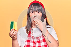 Young plus size woman wearing apron holding scourer covering mouth with hand, shocked and afraid for mistake